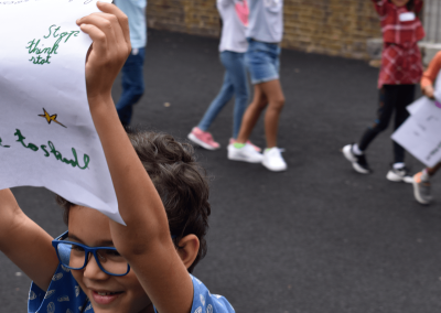 Children march in a playground holding banners showing what they care about as part of Summer Camps with Big Ideas for Westminster City Council.