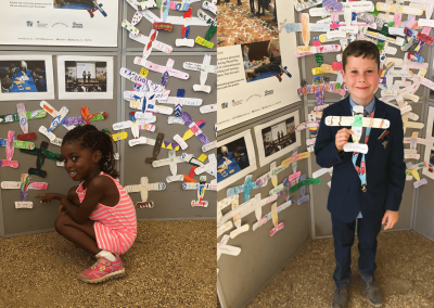 Two young Remember RAF100 participants stand in front of a display of hundreds of colourful paper planes holding their own paper plane submissions to the commemoration project.